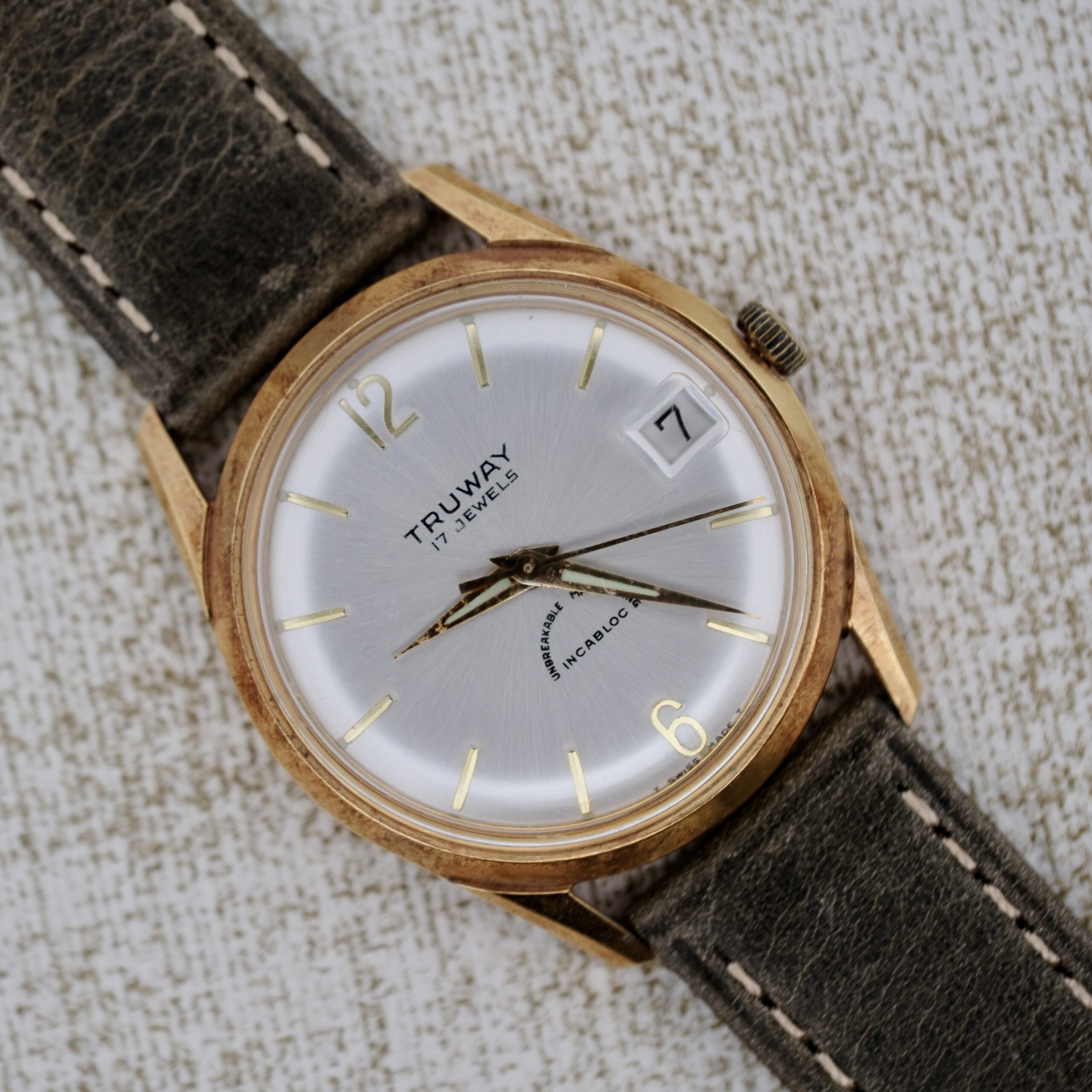 A Truway 17 Jewels Date Incabloc wristwatch with a silver dial, gold case, black hands, date window at 3 o’clock, and a worn dark leather strap is displayed on a light textured surface.