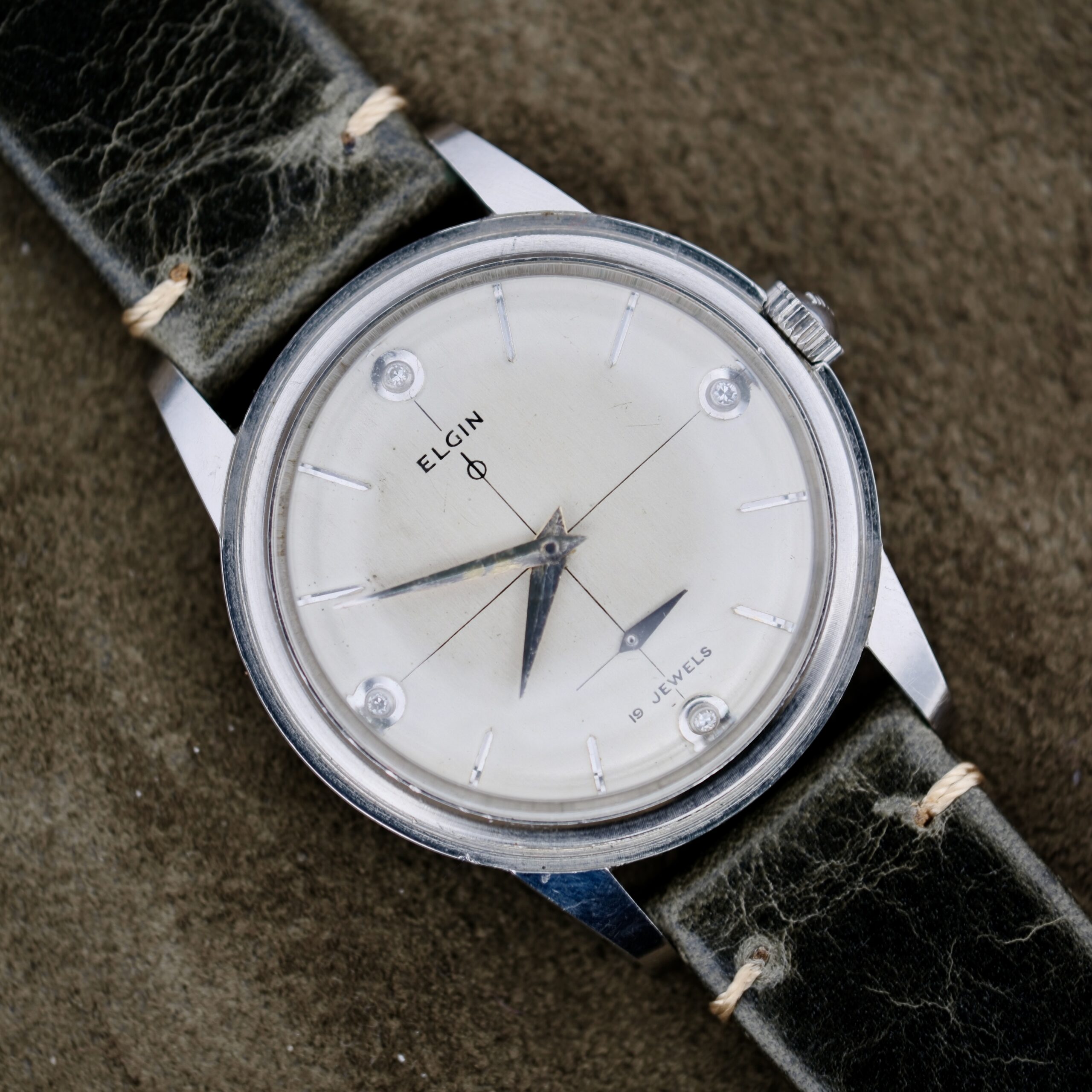The Elgin 19 Jewels Manual Wind Diamond Indicies features a vintage style with diamond markers at 12, 3, 6, and 9 on a textured brown dial, silver hands, and a black leather strap.
