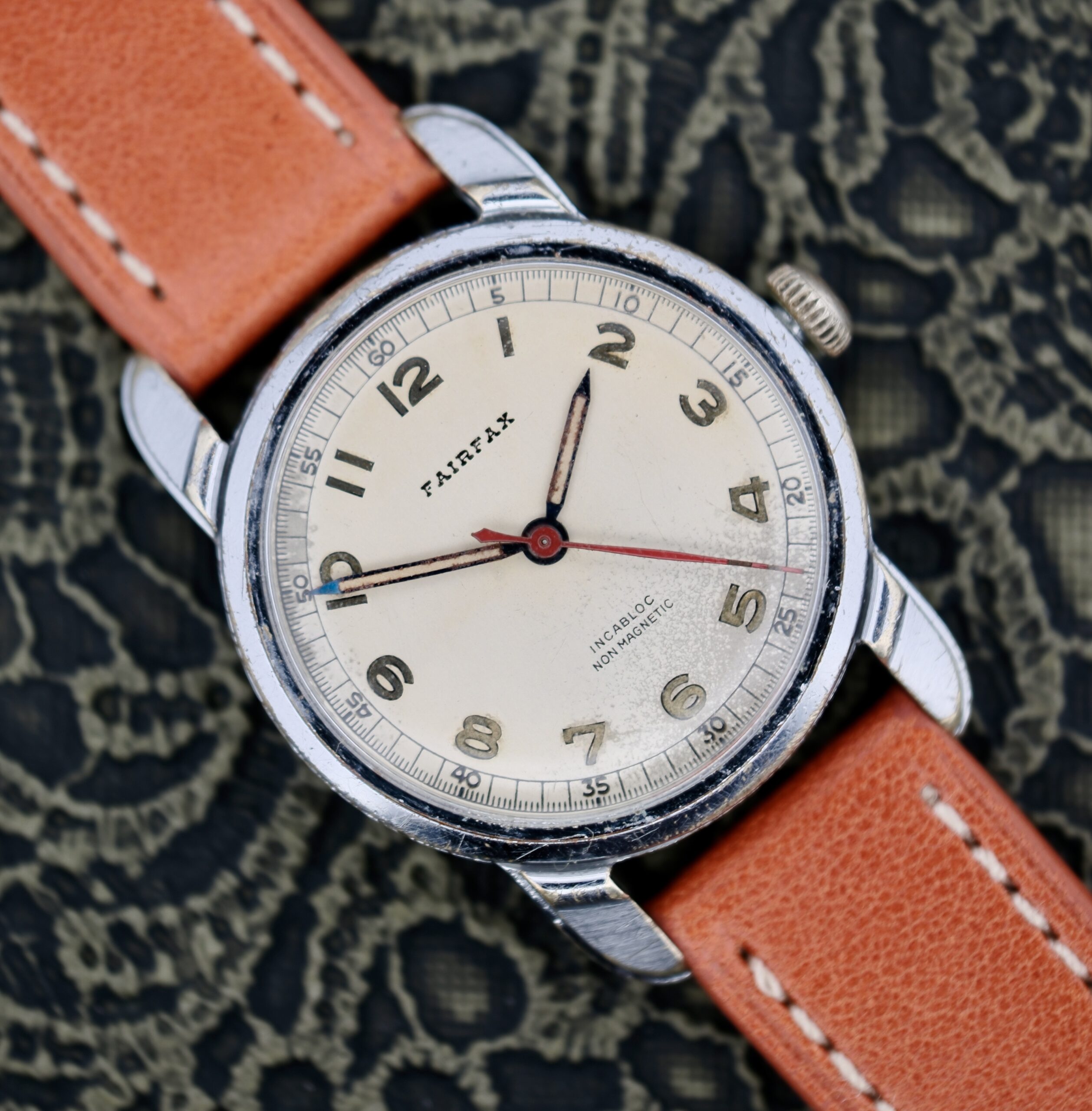 A 1940s Fairfax Military Incabloc Manual Wind wristwatch with a round cream dial, black numerals, silver casing, brown leather strap, and red seconds hand, displayed on a patterned black surface.