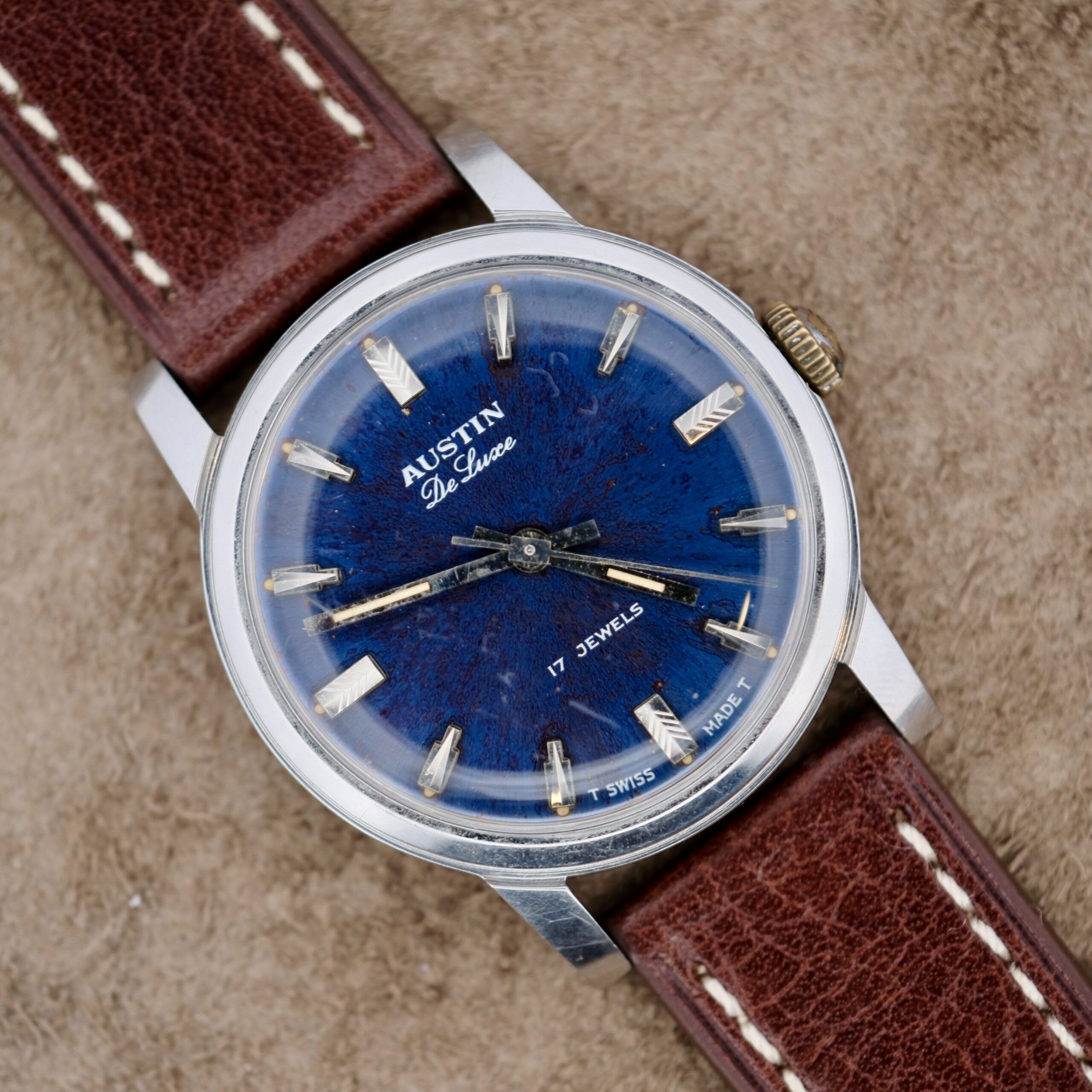 An Austin Deluxe 17 Jewels wristwatch with a blue patina dial, silver hour markers, and a brown leather strap with white stitching is displayed on a tan textured surface.