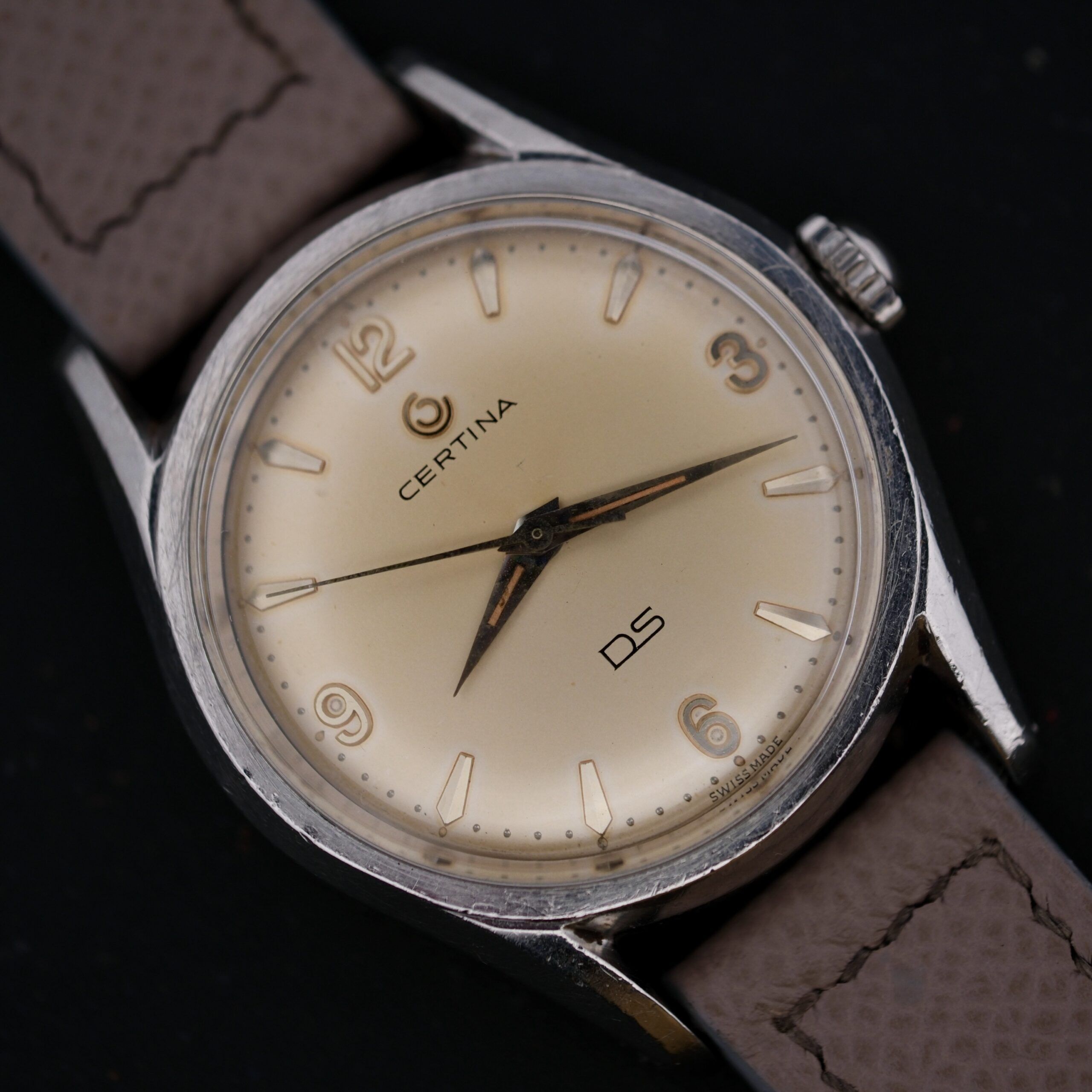 Close-up of a Certina DS 1st Gen Cal. 25-36 in stainless steel with a beige face, brown leather strap, and Arabic numerals at 12, 3, 6, and 9 oclock. Silver hour and minute hands enhance the design against a dark backdrop.