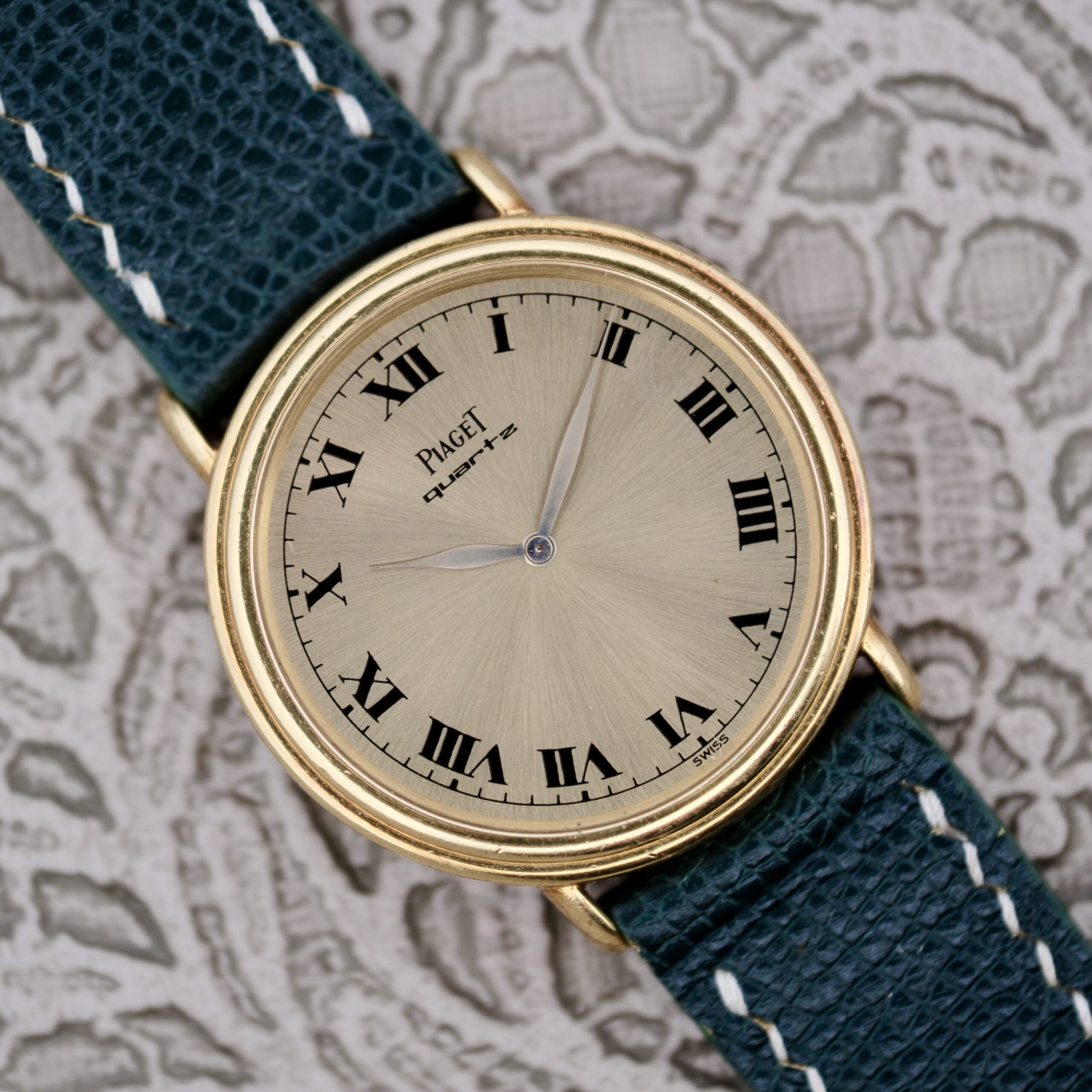 The Piaget Quartz 7632 18k Gold Champagne Dial is a classic round watch with a beige face, black Roman numerals, thin black hands, and a black leather strap featuring white stitching. The brand name graces the elegantly textured surface of the dial.