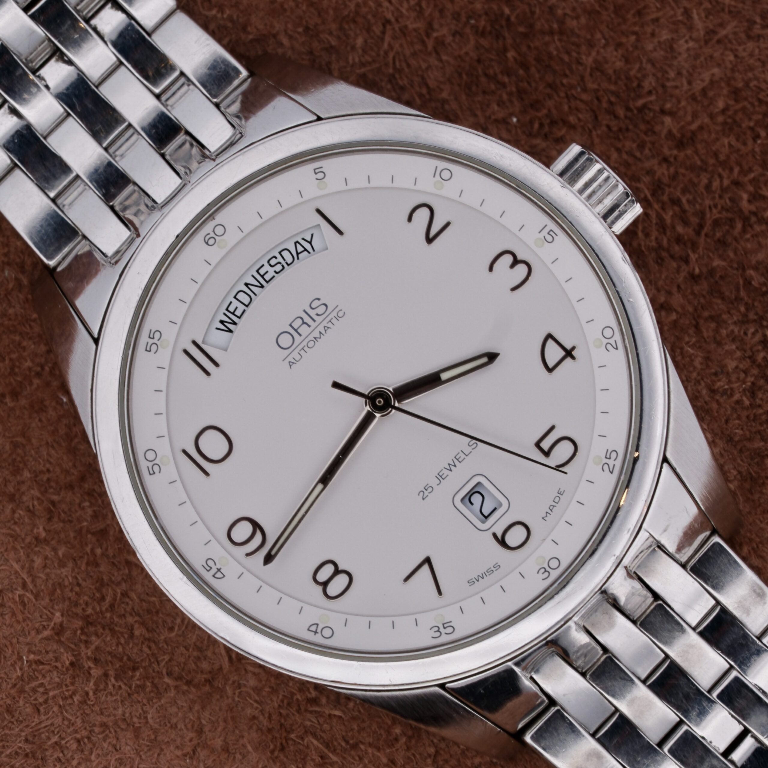 An Oris XXL Full Day Automatic silver wristwatch sits on a brown textured surface, featuring a metal band. The watch displays Wednesday, the date 28, and the time 10:10. The brand name Oris appears on its white dial.