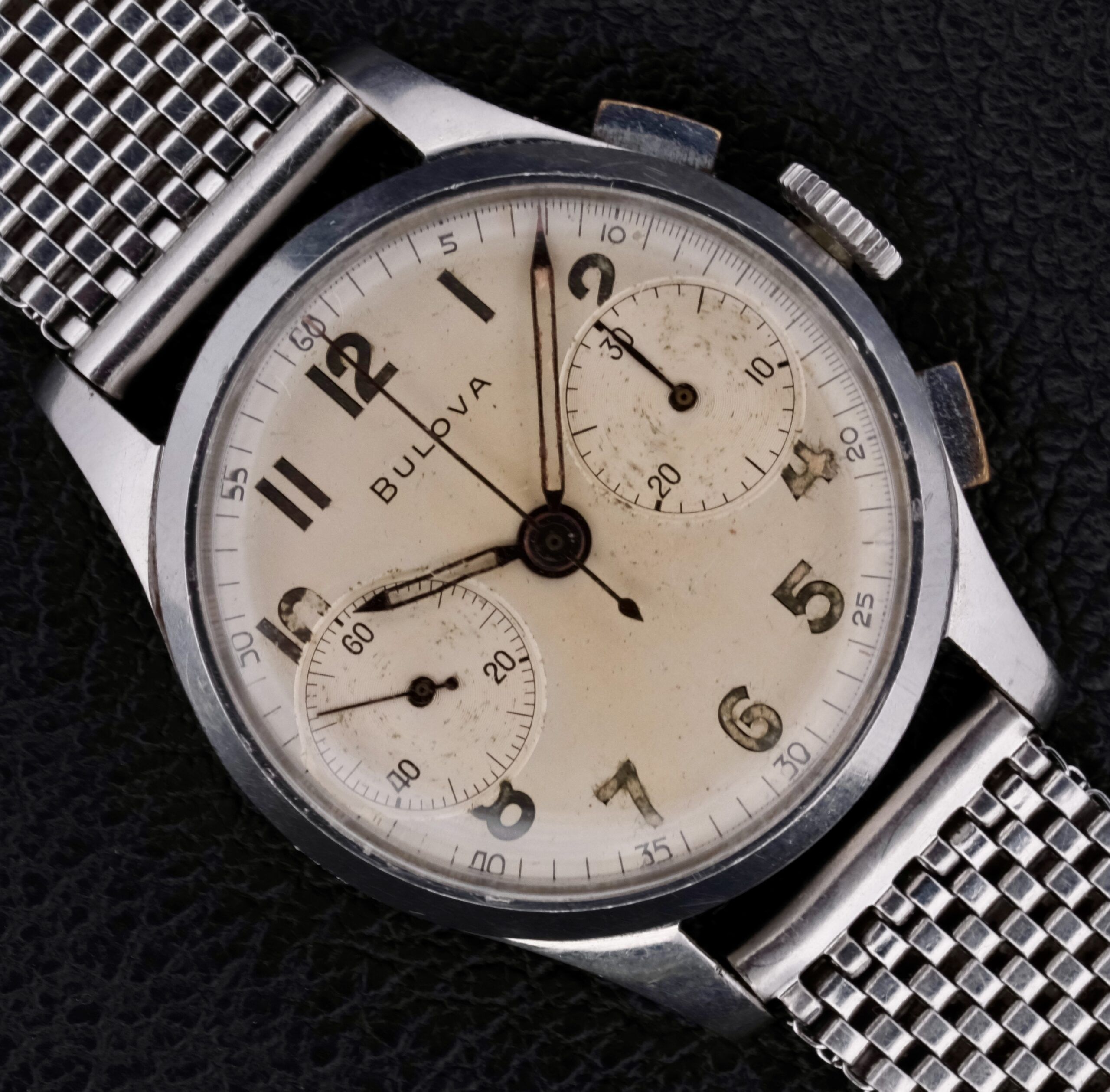 A Bulova Chronograph 13AK Valjoux 23 Stainless Steel watch with a silver mesh band rests on a black textured surface, featuring a cream-colored dial with three subdials, silver hands, and Arabic numerals.