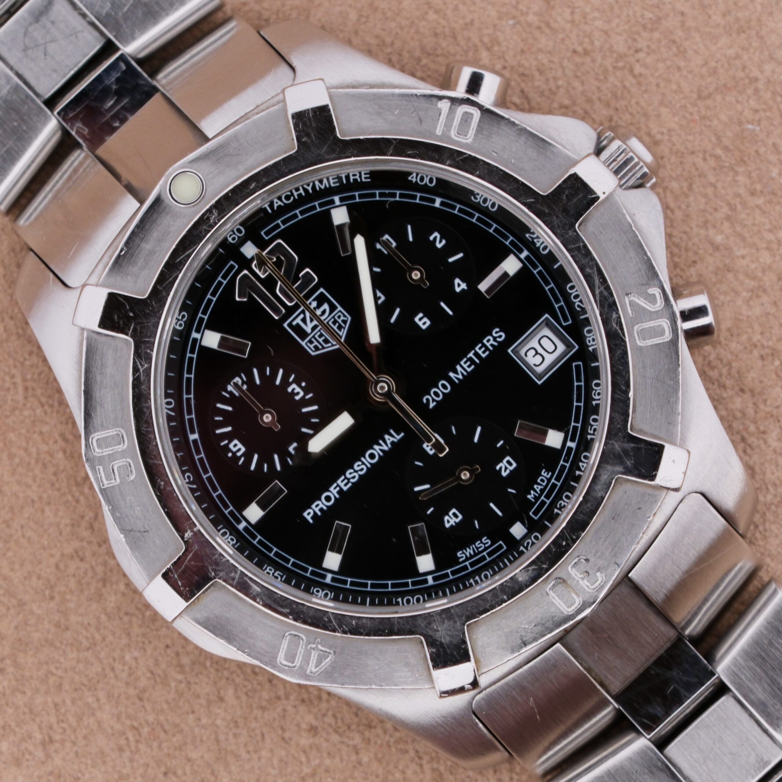 Close-up of a silver stainless steel Tag Heuer 2000 Professional Chronograph, showcasing its black dial with three sub-dials, date window, and tachymeter bezel. The watch face reads 200 meters and Professional, set against a light brown surface.