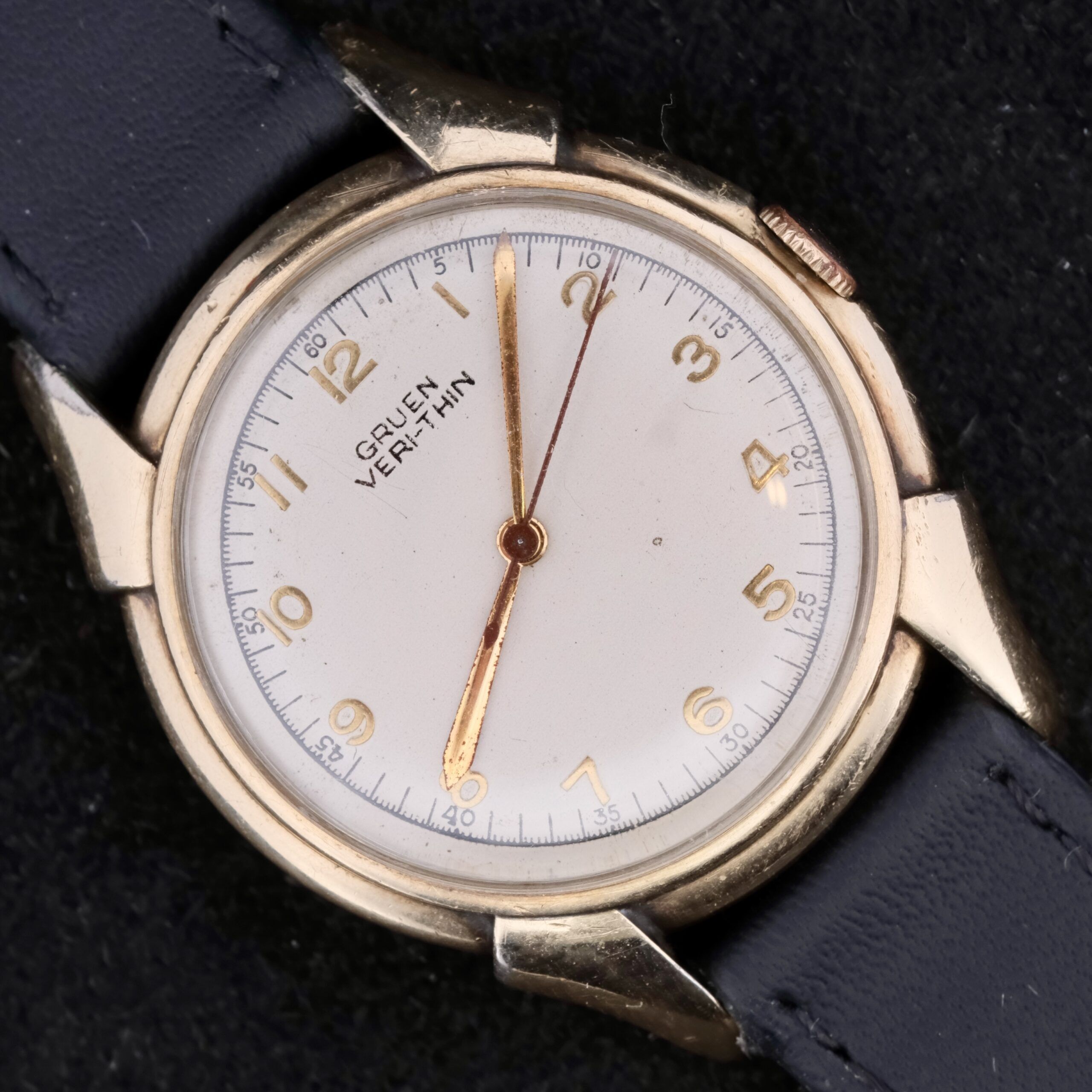The Gruen Veri-Thin Fancy Lugs vintage wristwatch showcases a round white face with gold hour markers, sleek hands, and an elegant gold-toned case. Paired with a black leather strap, it stands out against a sophisticated black background for a classic design.