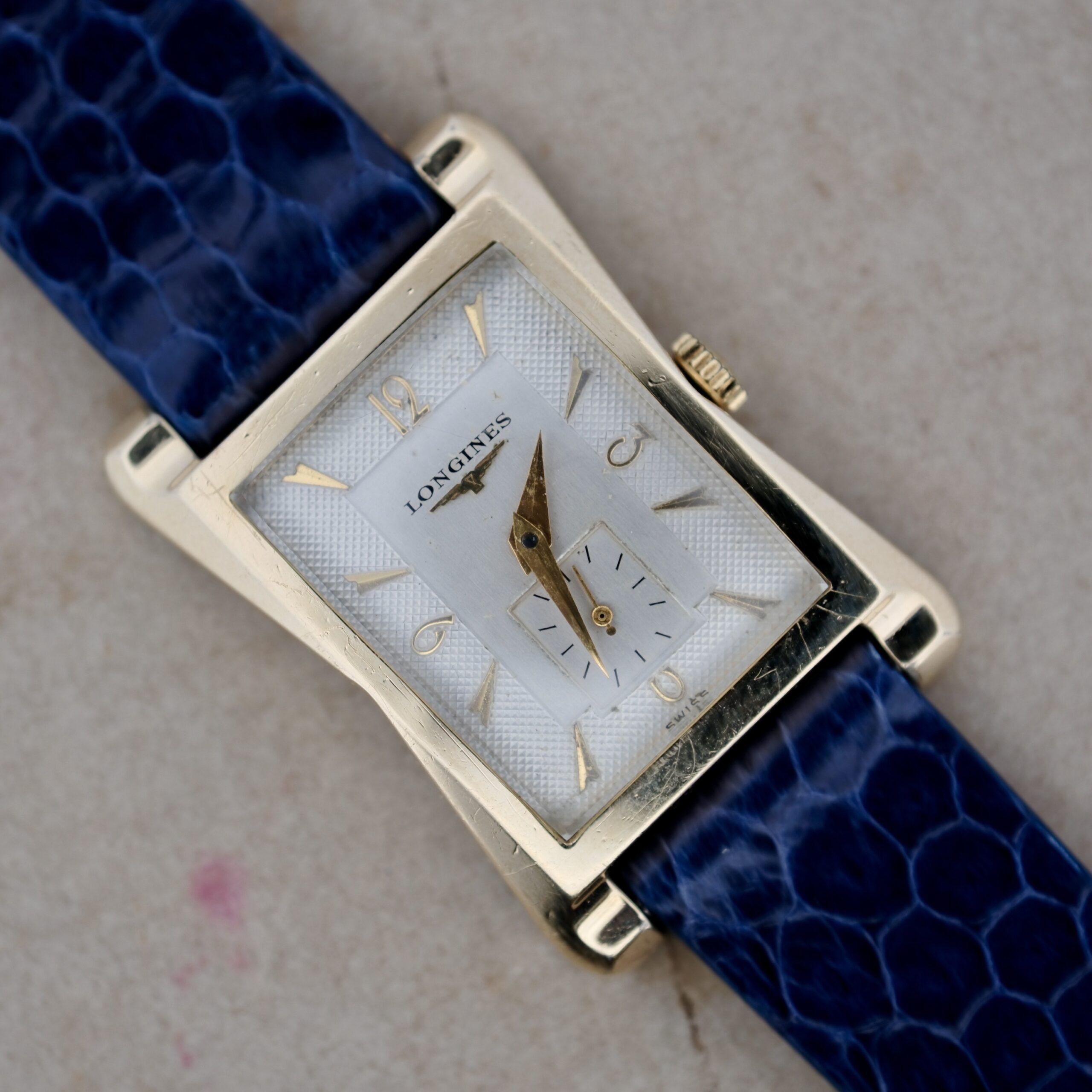 The 1950s Longines “Hour Glass” Case watch features a rectangular gold-toned design, white dial with black minute and hour hands, gold hour markers, a small seconds sub-dial at 6 oclock, and is paired with a blue textured strap.