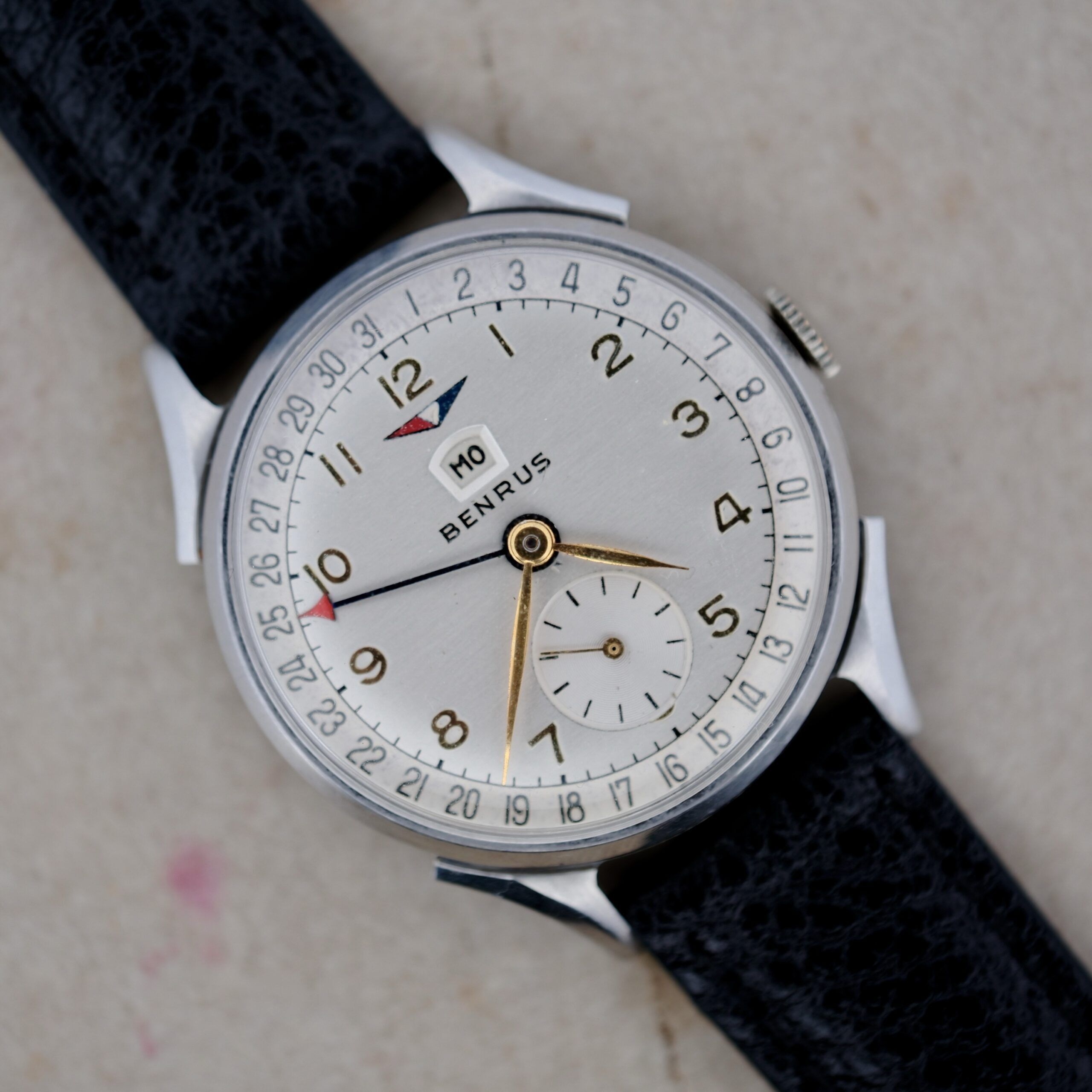 Close-up of a Benrus Triple Calendar In Steel wristwatch with a black leather strap, showcasing a white dial, silver case, gold hands, date window at 12 oclock, and small seconds subdial above 6 oclock, resting on a light surface.