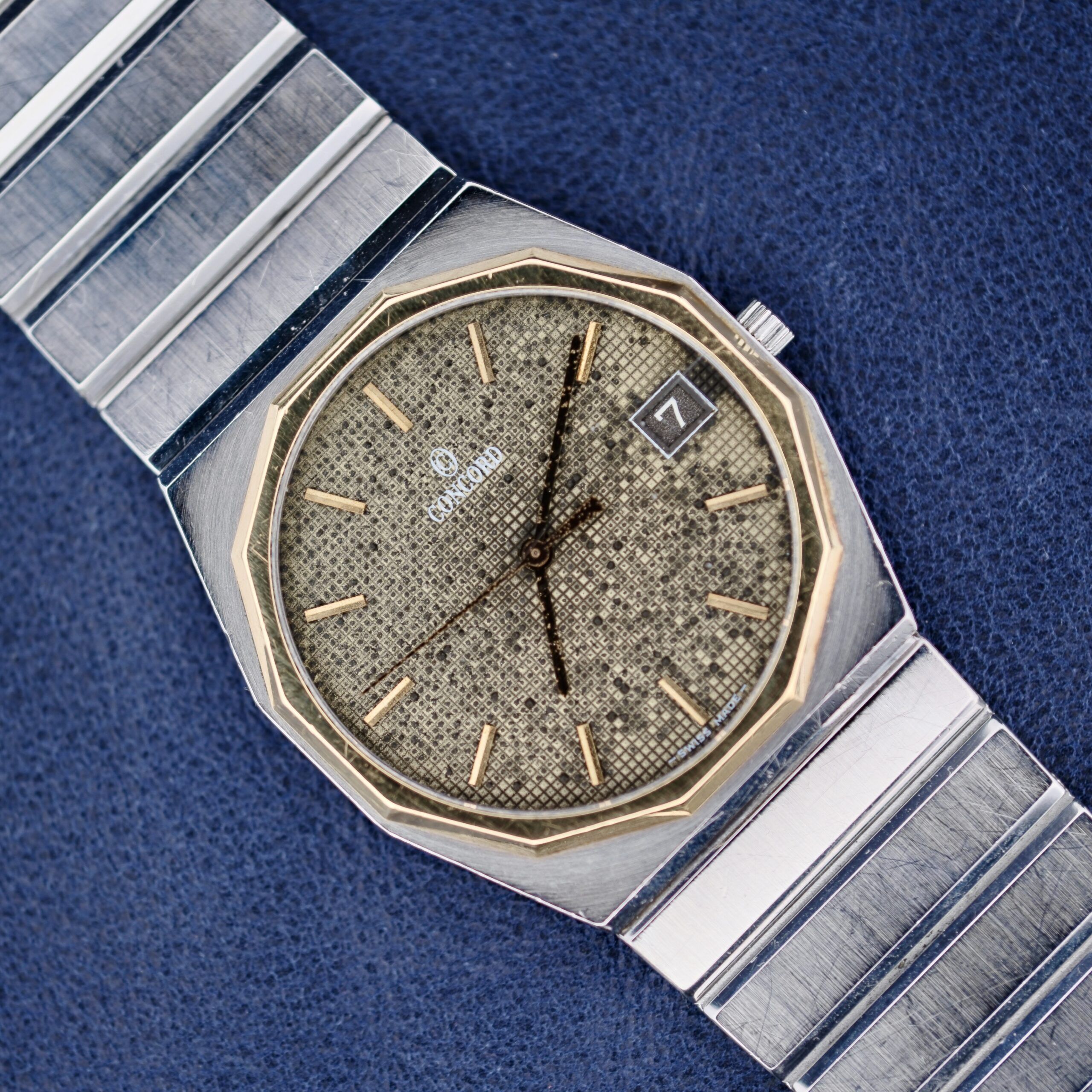 The Concord Mariner SG 18k Gold Bezel wristwatch showcases a textured gold-tone dial with gold hour markers and hands, complemented by a silver octagonal bezel. It features a date display at three oclock and a metal band with alternating shiny and matte links.