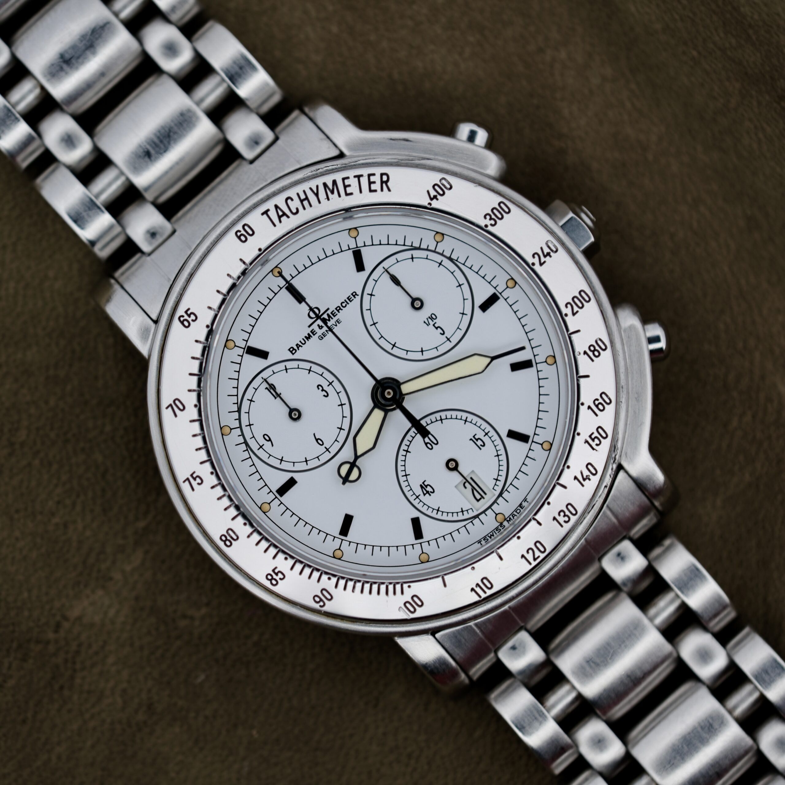 The Baume Mercier Formula S Chronograph, a silver wristwatch with a tachymeter bezel and three subdials, is displayed against a dark fabric backdrop. It features a white face, silver hour markers, luminous hands, and polished links on its metal bracelet.