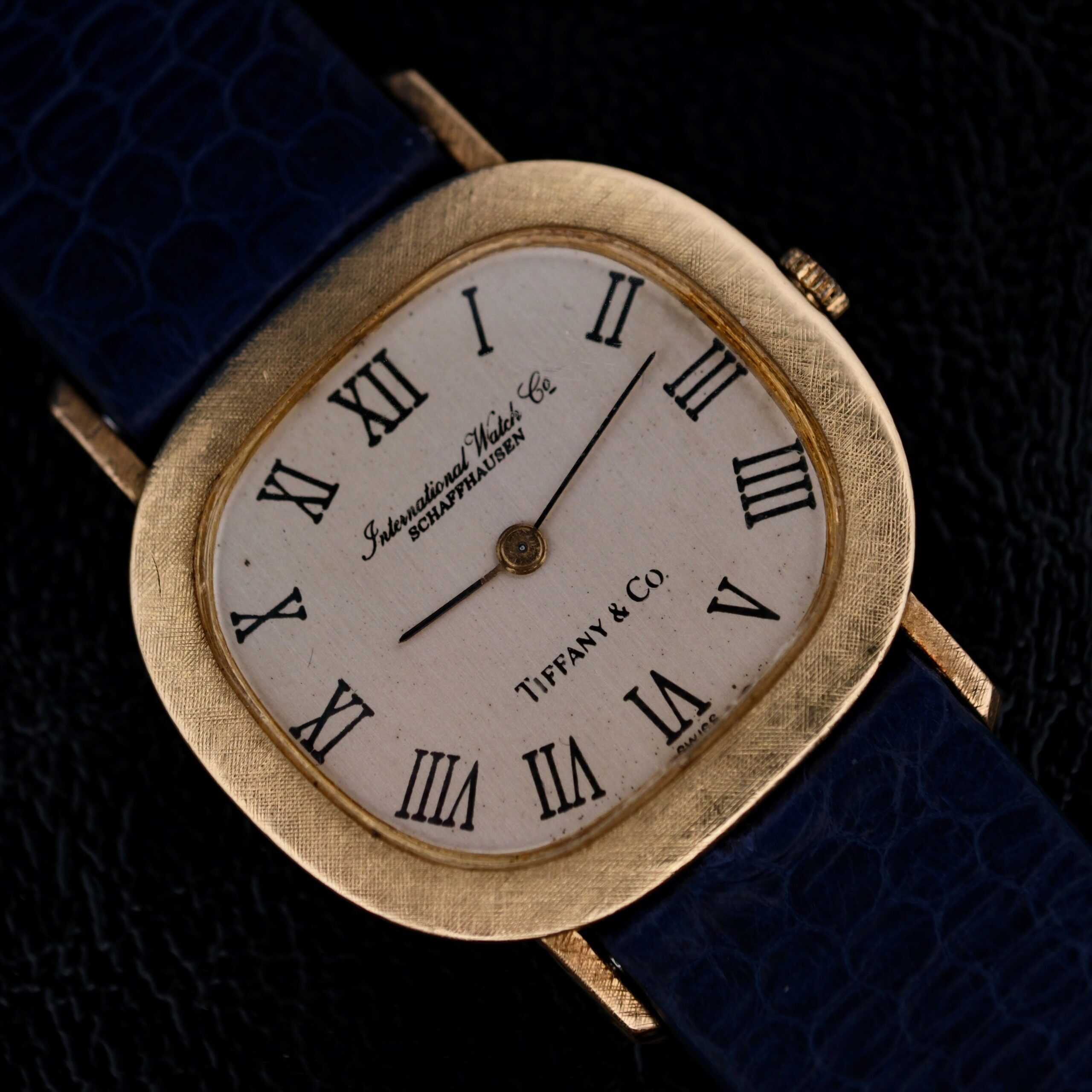 An IWC International Watch Co. men’s 18k gold wristwatch features a square case, white dial with black Roman numerals and hands, Tiffany & Co. stamp, textured bezel, and a dark blue leather strap.