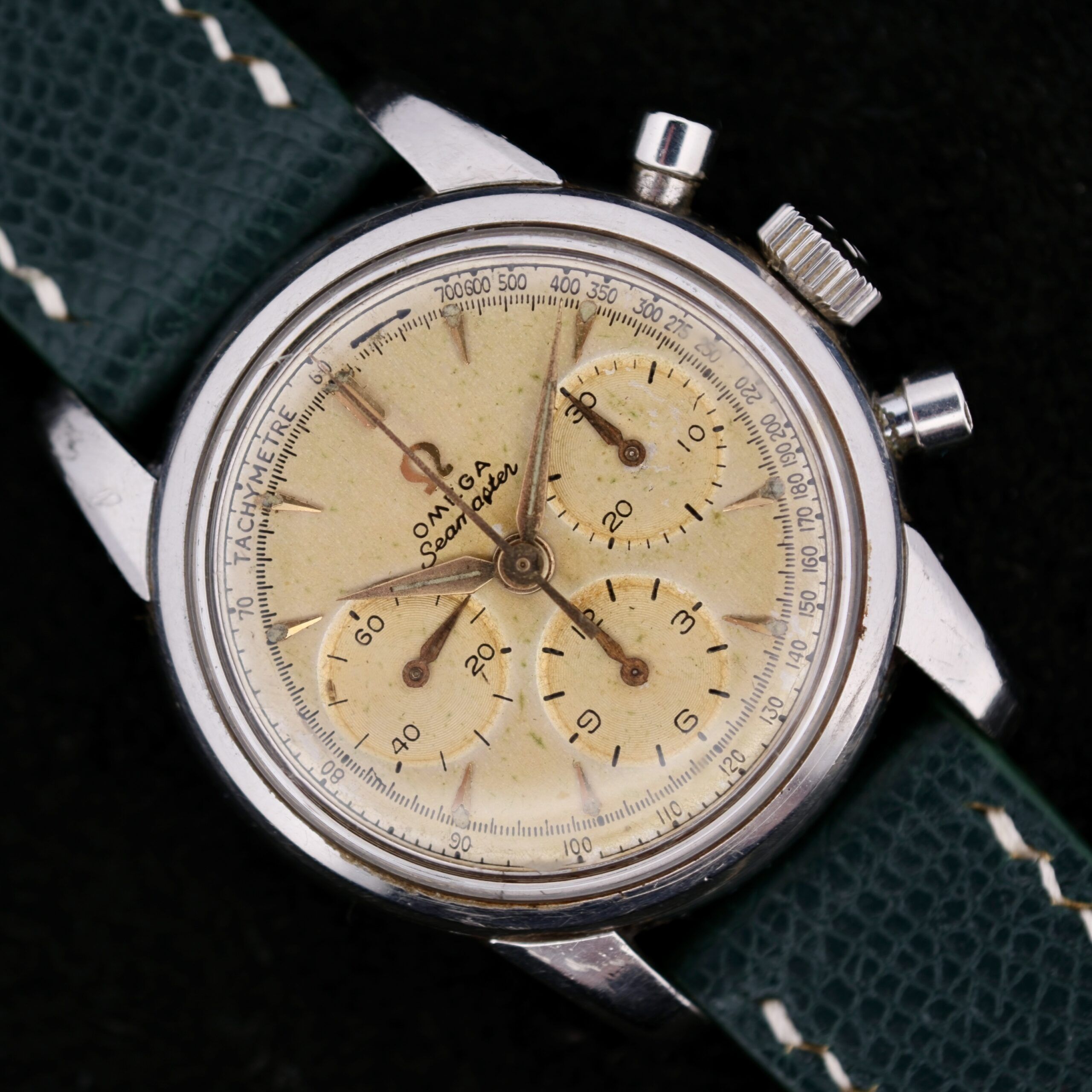 The Omega Seamaster Chronograph KO 2907 is a vintage watch featuring a cream dial, silver case, and three subdials. It has a tachymeter scale on the outer rim and comes with a textured green leather strap and thin metallic hands.