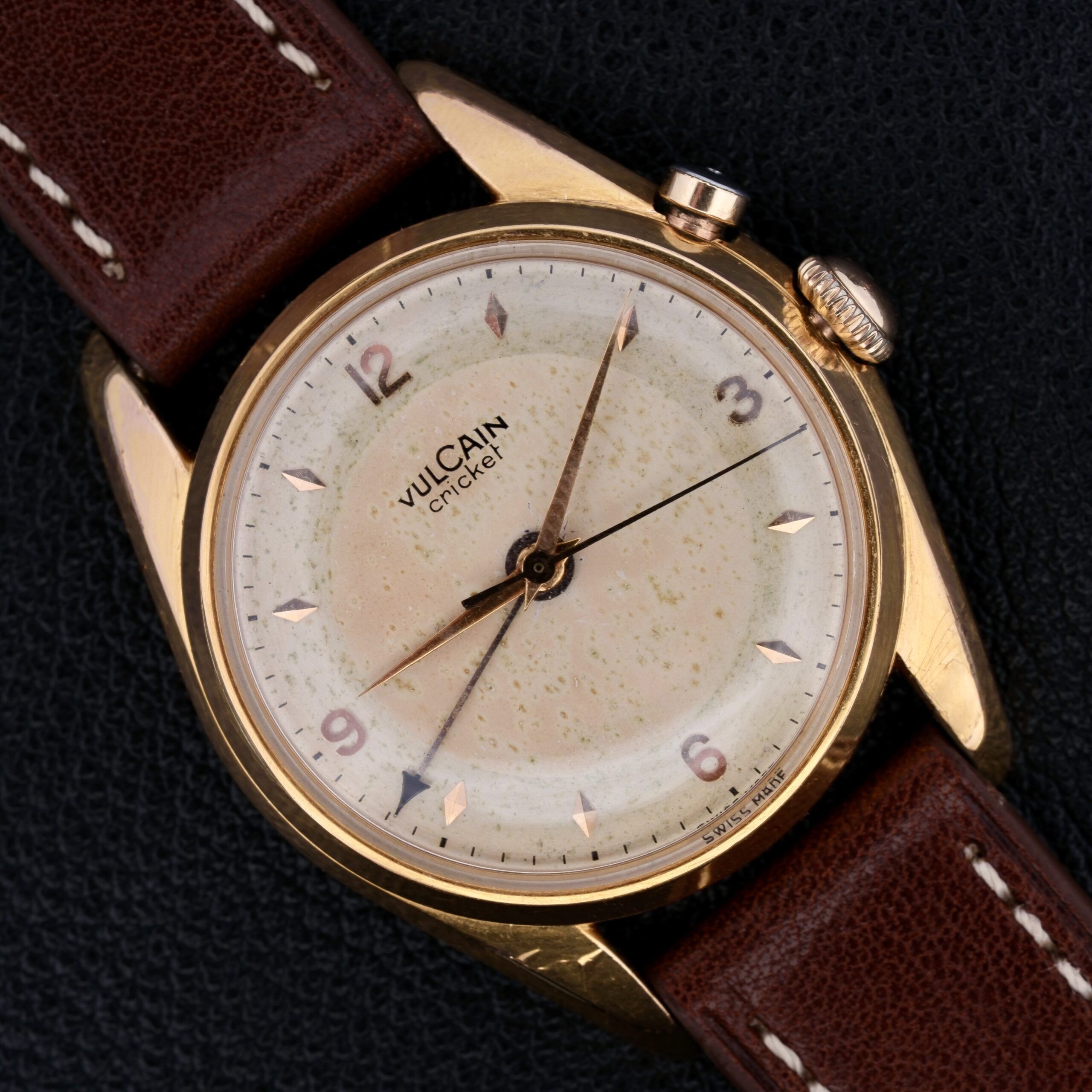 A vintage Vulcain Cricket 17 Jewels wristwatch featuring a gold casing, cream dial, and brown leather strap. It displays Arabic numerals at 9, 12, and 3 with gold hour markers and hands on a dark textured background.